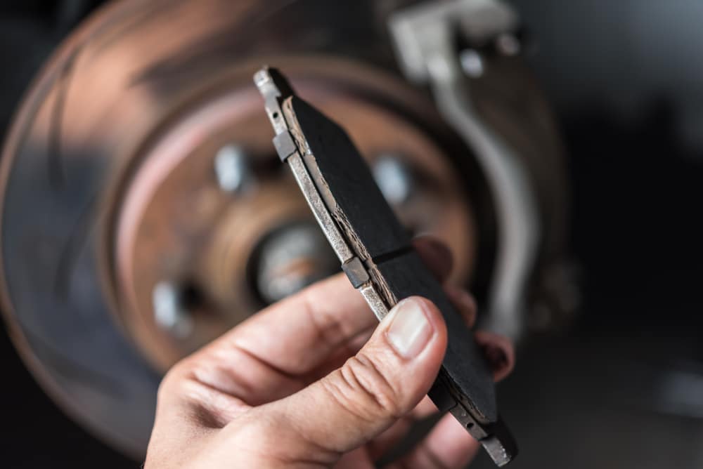 Brake pad inspection by Done With Care Auto Repair in Merriam, KS. Image showing a hand holding a worn brake pad with a brake rotor visible in the background, highlighting the importance of regular brake pad inspections for vehicle safety.