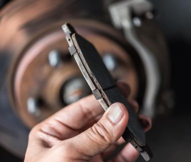 Brake pad inspection by Done With Care Auto Repair in Merriam, KS. Image showing a hand holding a worn brake pad with a brake rotor visible in the background, highlighting the importance of regular brake pad inspections for vehicle safety.